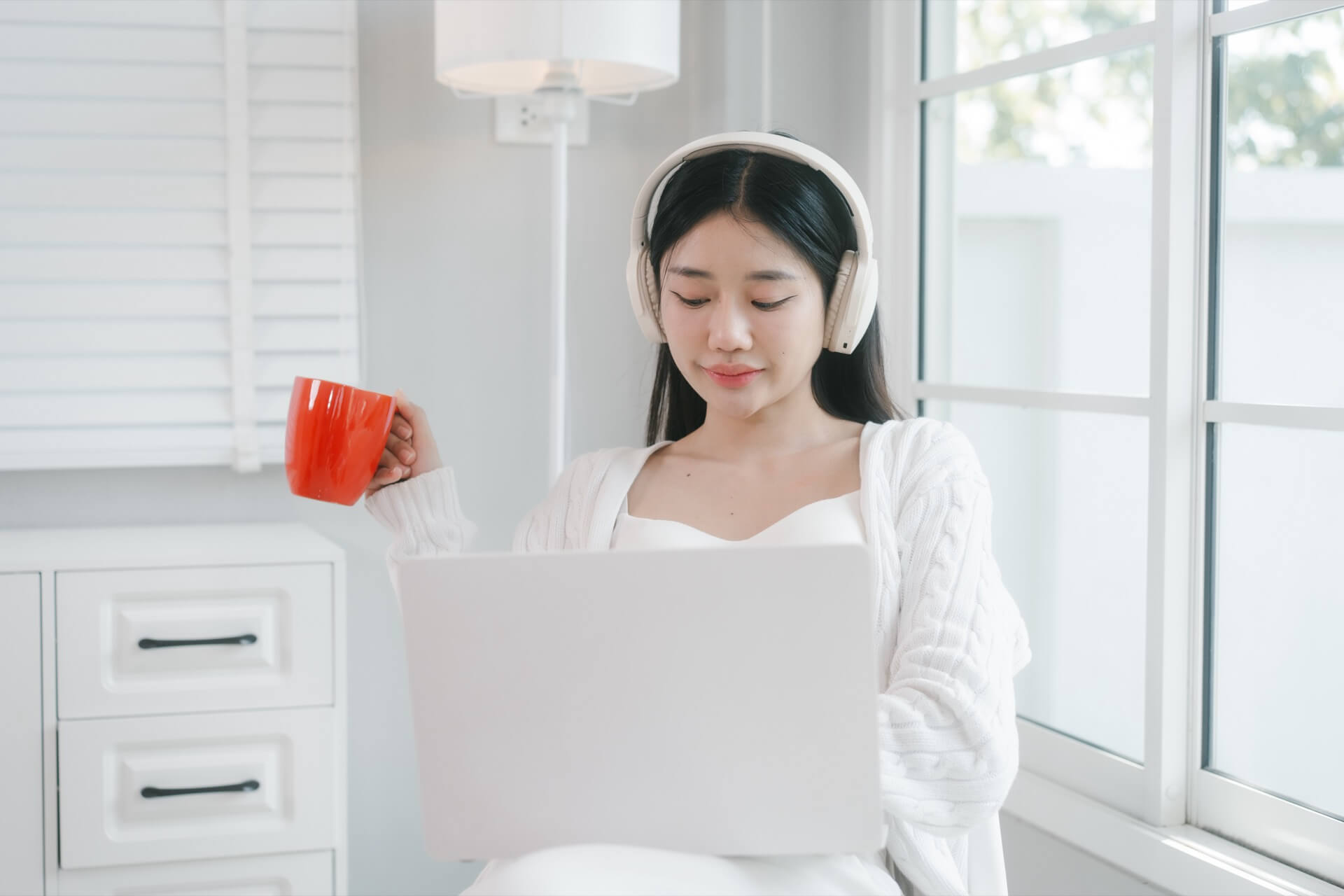 明るい窓辺でヘッドホンを着け、ノートパソコンで作業する女性
