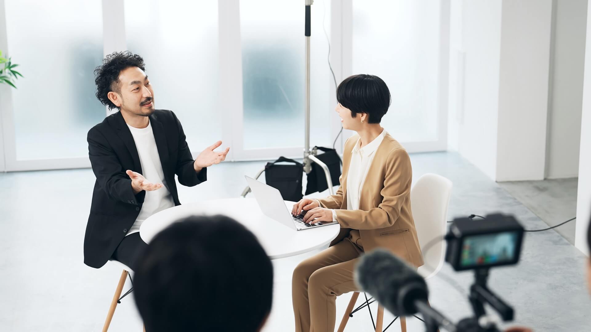 明るいスタジオでカメラとマイクを構え、男性へのインタビューを収録する様子