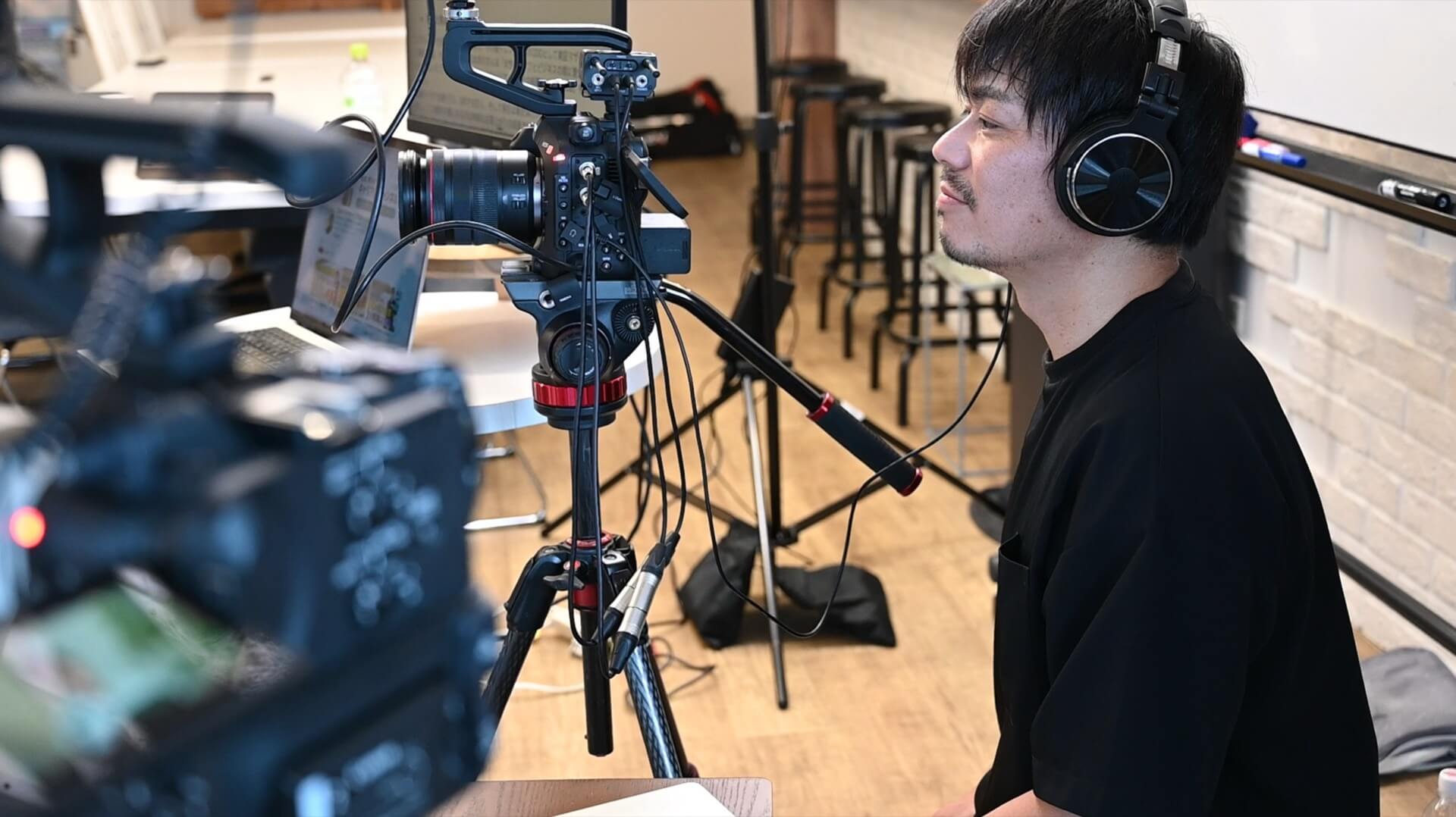アクエリアス・ムービーの撮影現場で、三脚のシネマカメラの横でヘッドホンを付けて音声を確認するスタッフ