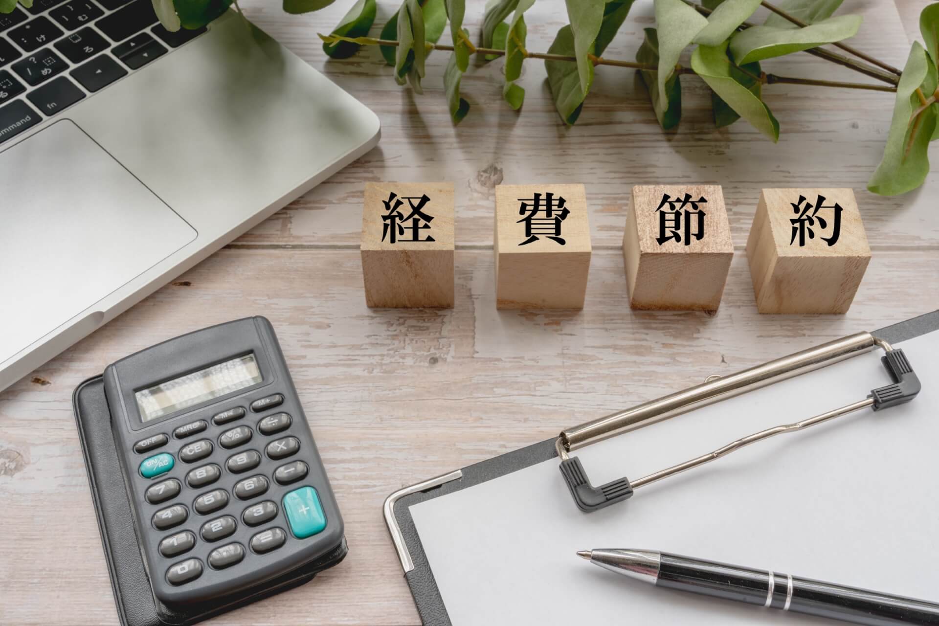 「経費節約」の文字ブロックと電卓が置かれたデスク