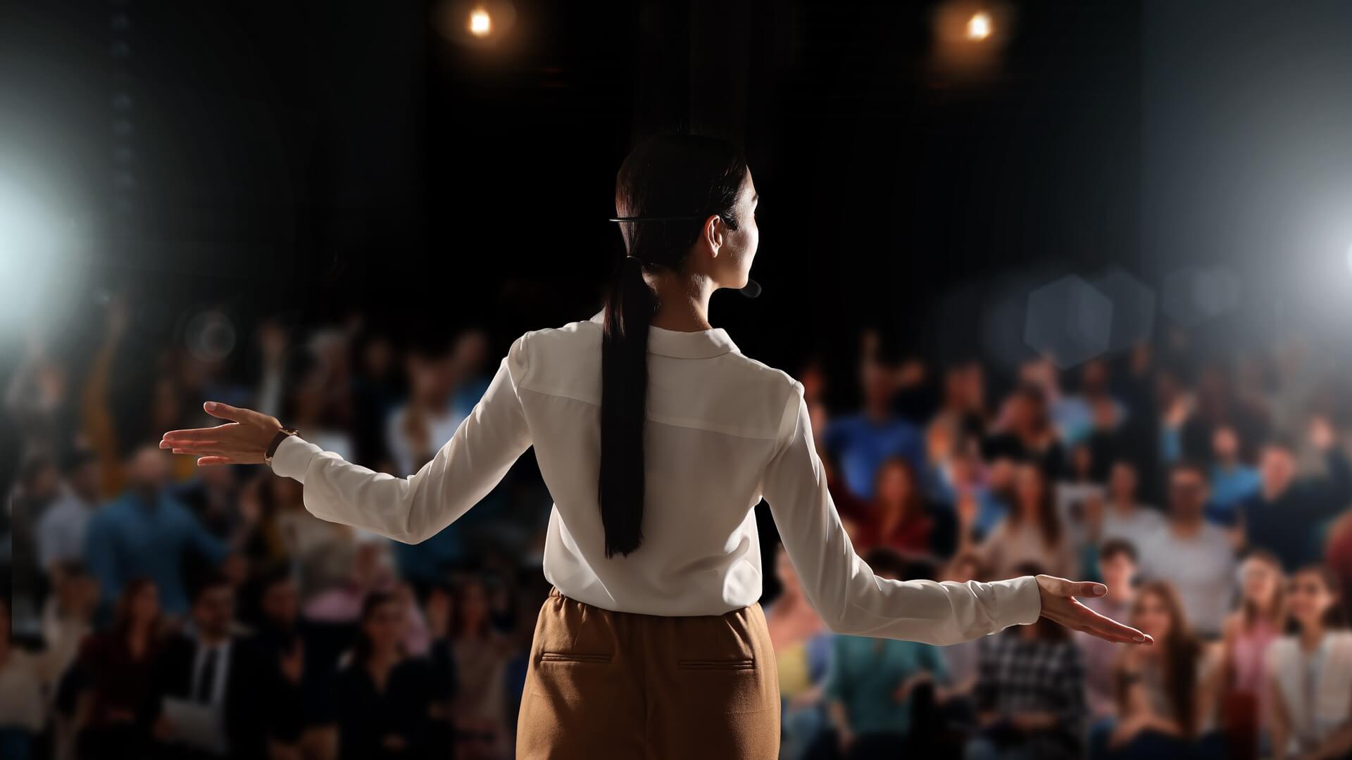 スポットライトの当たるステージで、満席の会場に向かって両手を広げる登壇者の後ろ姿