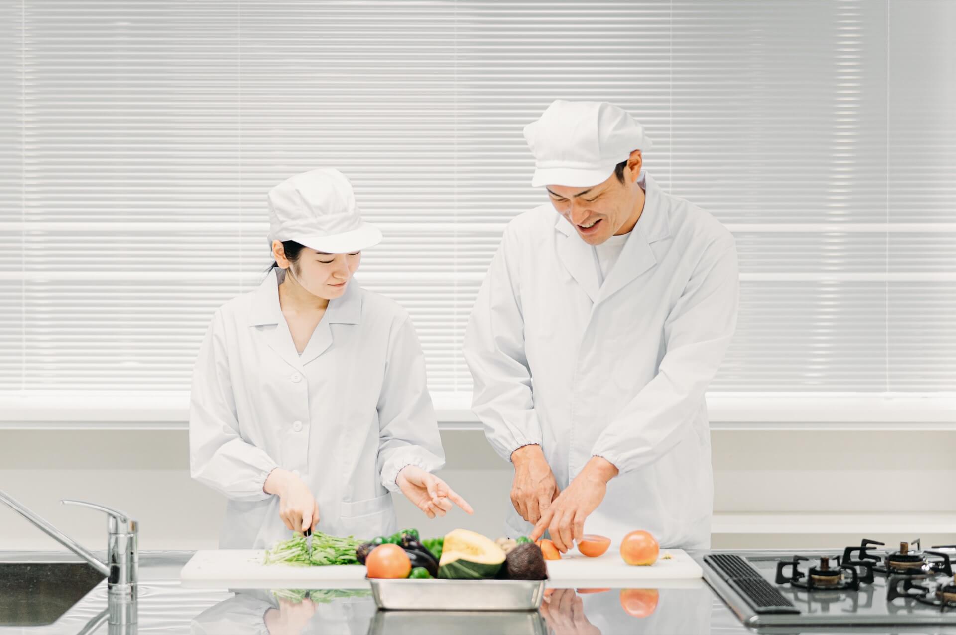 調理場で、先輩の調理員が新人に野菜の切り方を教えている様子