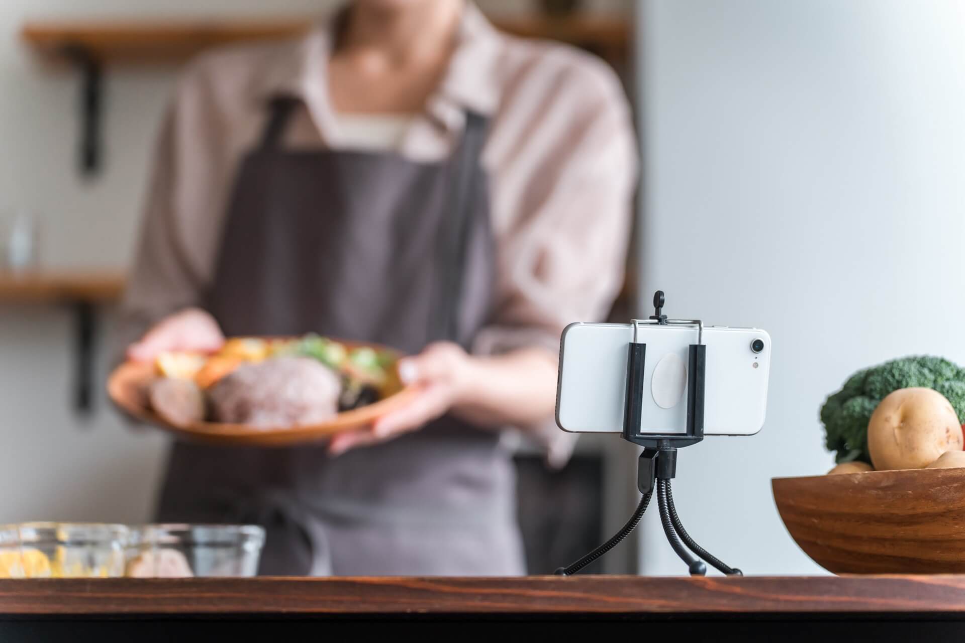 キッチンで、三脚に固定したスマートフォンで料理の手順を撮影する女性