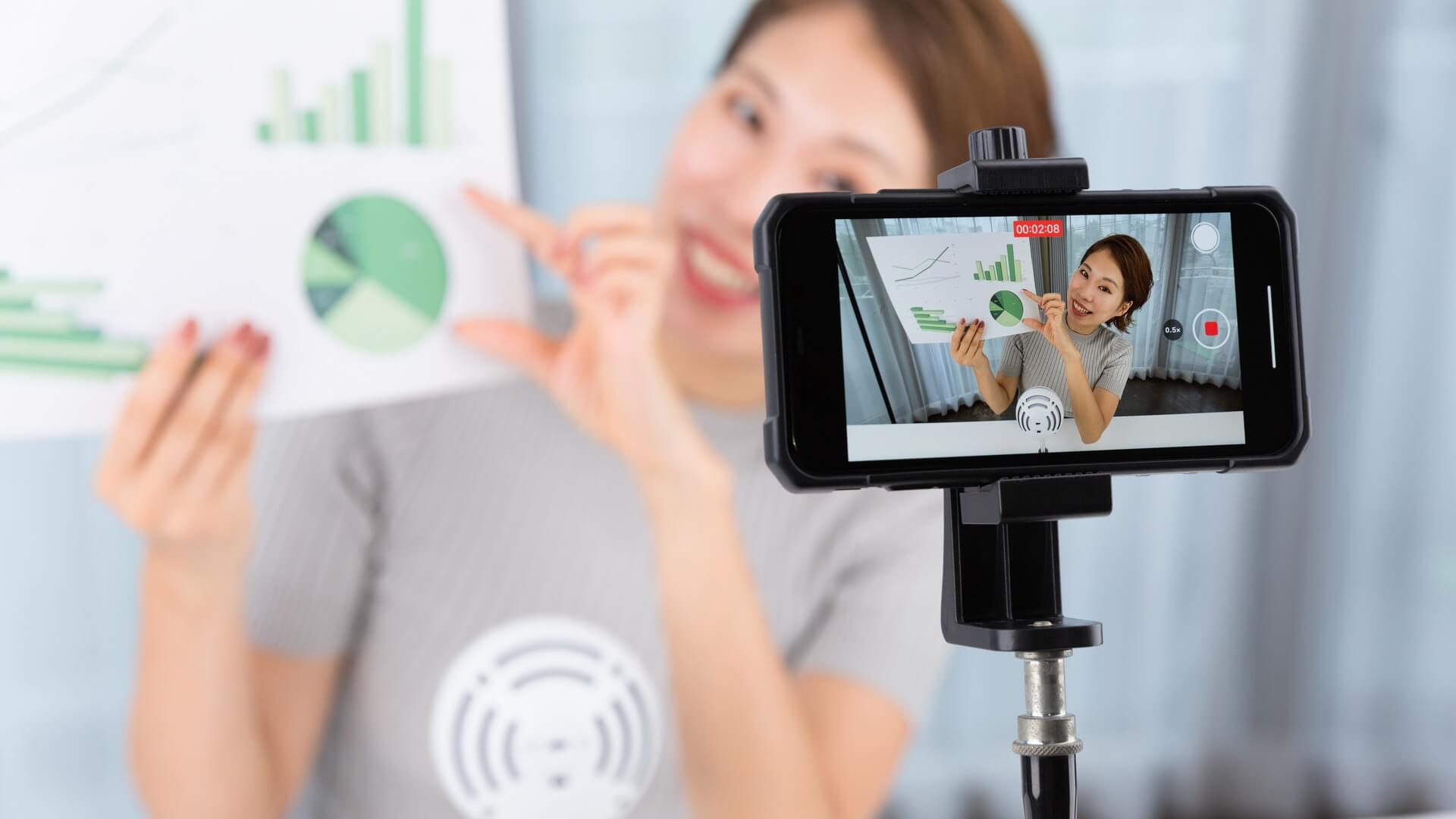 グラフを手に、スマートフォンで動画を撮影しながら説明する女性