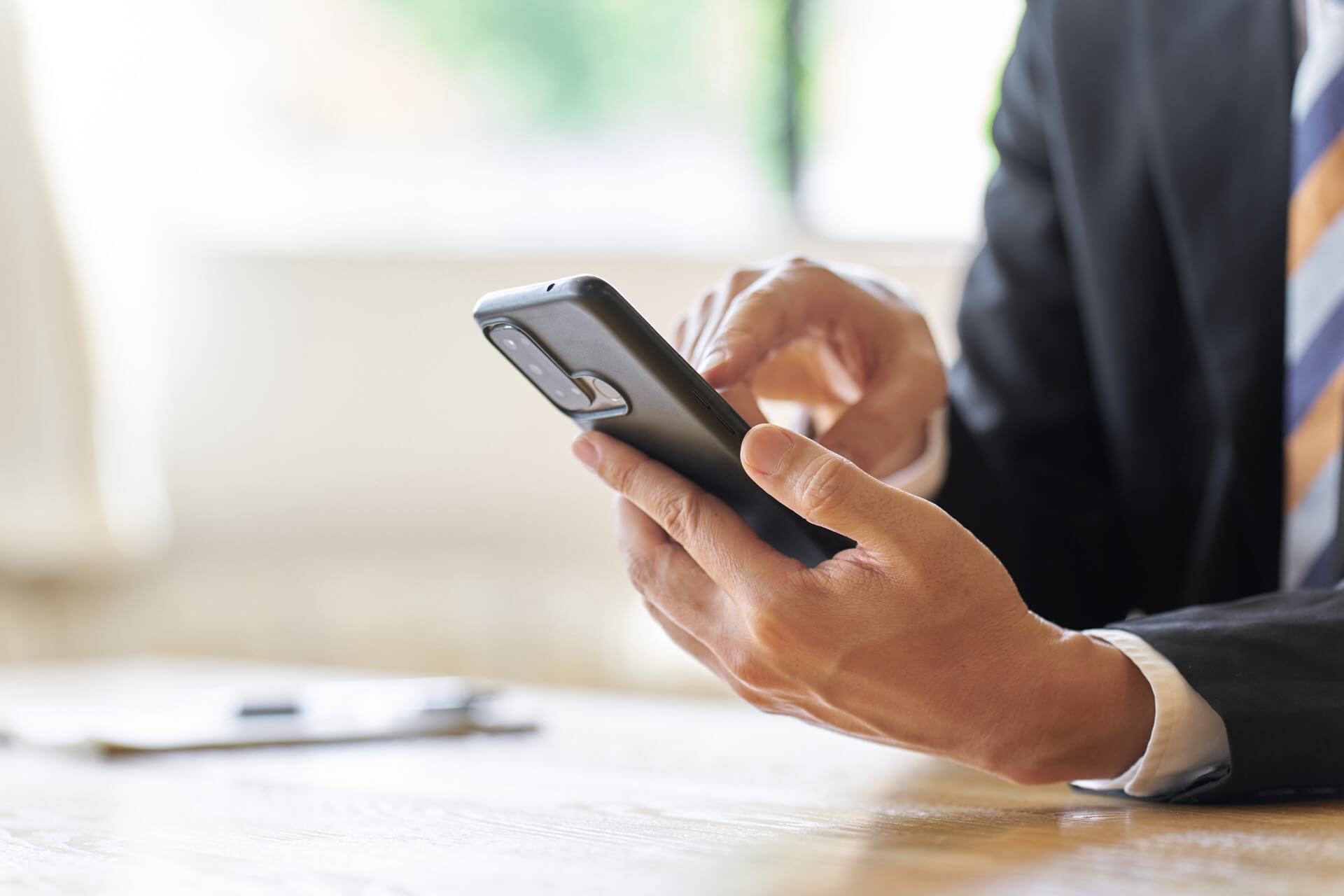 自然光の入るデスクで、スマートフォンを操作するスーツ姿のビジネスパーソンの手元